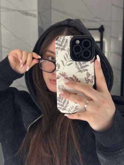 A partial reflection of an individual, likely female, is visible in a mirror, indicative of a selfie being taken indoors. The subject has long, dark brown hair and is wearing a dark, possibly charcoal grey, zip-up hooded sweatshirt with the hood up. Her left hand is raised, holding and adjusting a pair of black-framed eyeglasses positioned across her face. Her right hand holds a smartphone, obscuring the lower half of her face. The smartphone is equipped with a triple-lens camera array and housed in a cream-colored case featuring a vertical ribbed pattern adorned with botanical illustrations of leaves, flowers, and butterflies in muted tones. Both hands display light-colored, oval-shaped manicured nails. A metallic ring with a patterned inlay is worn on the ring finger of the right hand. The visible portion of the subject's face shows a direct gaze from the left eye. The background reflection consists of light-colored, possibly grey, tiled walls. A dark, vertical fixture, potentially a towel rack, is visible in the upper right portion of the reflection. The specific location within the city of Prikubanskii, Russia, cannot be determined from the provided image.