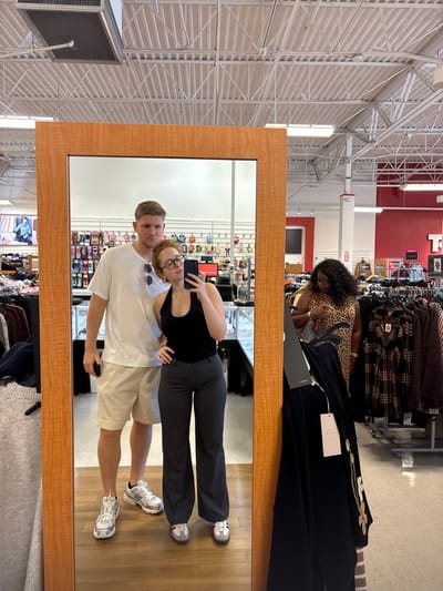 The image depicts the interior of a TJ Maxx retail store, located in West Windsor, United States. A large, vertically oriented mirror with a light-colored wooden frame dominates the foreground, reflecting two individuals posing for a mirror selfie.
The female in the reflection, positioned in front and slightly right of the mirror's center, has reddish-brown hair, wears glasses, a black halter top, dark gray wide-leg pants, and light-colored sneakers. She holds a dark-colored smartphone with her right hand, pointed towards the mirror to capture the image. A delicate necklace and a ring on her left hand are visible.
Behind and to the left of the female, a male is reflected. He has short blond hair, wears a white t-shirt with sunglasses hooked on the collar, light beige shorts, and white/silver athletic sneakers. He holds a phone in his right hand, positioned at his side. Both individuals are standing and looking towards the phone, posing for the photograph.
In the unreflected background, on the far right, a woman with dark, curly hair, wearing a leopard-print garment, is partially visible. She appears to be interacting with or observing items on a clothing rack, potentially holding a phone or merchandise.
The store environment features high, exposed white ceilings with metal trusses, ventilation ductwork, and fluorescent strip lighting. Merchandise includes racks of clothing, shelves displaying sunglasses or small accessories, and glass display cases likely containing jewelry. A distinctive red wall section with a white letter "T" (part of the "TJ Maxx" logo) is visible in the right background, along with a fire extinguisher mounted on a white support column. In the unreflected foreground, a gray ribbed garment is partially visible on the left, and a dark garment with two white price tags, one indicating "VITA MILANO," hangs on a display stand to the right of the mirror frame.