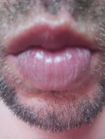 A close-up, slightly out-of-focus photograph shows the lower half of a person's face, specifically their mouth and the surrounding area with stubble. The lips are slightly parted, revealing a pinkish interior. The skin appears to have a light complexion with noticeable facial hair growth, indicating a male subject. The lighting is soft and diffused, creating a gentle illumination. The image context does not provide any information about the city of Таёжное, Russia, or any urban environment.