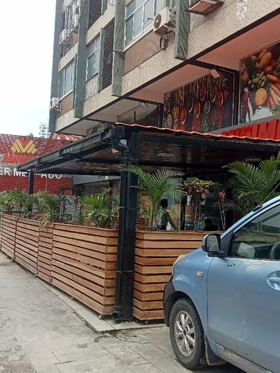 The image captures a street-level view of a commercial establishment in an urban setting, likely Luanda, Angola, given the visible Portuguese text. The scene is set during the daytime under an overcast sky.
In the background stands a multi-story building with a light-colored, possibly beige or brown, facade. Several large windows are visible, and air conditioning units are mounted on the exterior walls. Green mesh or netting is draped down parts of the building.
Extending from the building is a dark-colored, possibly metal, canopy providing shade over a patio or outdoor area. A black downspout is visible on one of the canopy's supporting pillars. Along the edge of this outdoor area, a series of light brown wooden slatted planters, some appearing to be on wheels, form a barrier from the sidewalk. These planters are filled with lush green foliage, including several small palm-like plants.
Behind the planters and under the canopy, a person is partially visible, seen from behind as a dark silhouette. Further to the left, behind the main canopy structure, a red-tiled roof with a yellow "M" logo is present, and partially visible text reads "ER MER ADO," strongly suggesting "SUPERMERCADO" (supermarket). On the building facade above the canopy, large photographic displays feature images of various spices in spoons and an assortment of fresh fruits and vegetables, indicating a food-related business. A small, unreadable sign with numbers like "1E-46" is affixed to the building above the spice display.
On the right side of the frame, the front quarter of a light blue compact sedan is parked at the curb, its front wheel and fender visible. No specific activity beyond typical urban presence is clearly depicted.