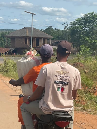 Two men are riding a red motorcycle on a dirt road in Ikoga, Nigeria. The driver is wearing a brown baseball cap, a light beige t-shirt with an "Aeronautica Fashion F/TF-1978 Homme London" logo on the back, and gray pants. The passenger is wearing a purple baseball cap and an orange shirt. They are carrying a large white bag tied with red and white fabric. In the background is a brown building with a metal roof and a utility pole. The environment is lush with greenery and trees, and the sky is blue with scattered clouds.