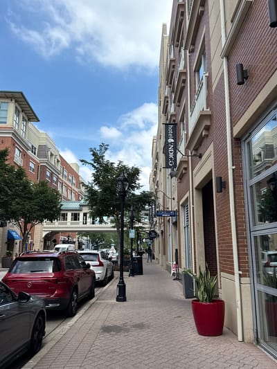 The image depicts a street scene in Naperville, United States of America. On the right side of the frame, a brick building with cream-colored accents and multiple windows lines the sidewalk. A sign for "hotel INDIGO" is visible on the building. A large red planter containing a snake plant sits on the sidewalk in front of the building. The sidewalk is paved with interlocking bricks. On the left side of the frame, a row of cars, including a red SUV and a gray car, are parked along the street. Further down the street, more buildings and trees are visible, as well as a pedestrian bridge connecting two buildings. The sky is blue with scattered clouds.