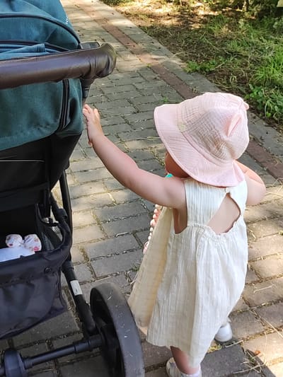 The image shows a young child wearing a light-colored, textured dress and a pink sun hat. The child is standing on a paved walkway next to a teal stroller. The child's right arm is extended towards the stroller's handle, and their left hand is also near the stroller. Inside the stroller's basket, a white item with a floral pattern is visible.
The scene appears to be outdoors, with grass and foliage visible in the background. The lighting suggests it is daytime, possibly late afternoon, with sunlight casting shadows. The overall mood is peaceful and domestic. The setting is a walkway made of rectangular pavers, suggesting a park, a garden, or a residential area.
No visible text is present in the image. The location context is Квасоўскі сельскі Савет, Belarus.