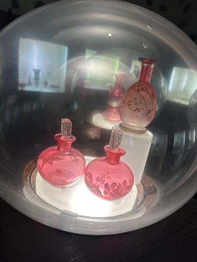 The image shows three pink glass perfume bottles with stoppers, arranged inside a large glass dome. Two of the bottles are in the foreground, and a third, taller bottle is visible behind them. The bottles appear to be antique, with decorative etching on the glass. The dome is placed on a reflective surface, which amplifies the light and reflections. In the background, there are blurred reflections of a room with furniture and possibly other decorative objects. The scene is lit from within the dome, creating a soft, ethereal glow. The image does not provide any information about a specific location in Trakai, Lithuania.