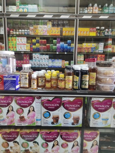 The image depicts a display of various health and wellness products within a retail store. The shelves are filled with boxes and bottles of what appear to be supplements, traditional medicines, and beverages. Prominently featured are multiple boxes of "Prenagen mommy" and "Prenagen lactamom" powder, aimed at pregnant and breastfeeding mothers, respectively. These boxes display images of women and nutritional information.
Other visible products include bottles of "Madu Pahit Asli" (Bitter Honey) and "Madu An-Nur Syifa" (Honey of Nur Syifa), suggesting a focus on herbal remedies and natural health products. There are also smaller bottles of various remedies or supplements, and some larger plastic containers filled with clear liquids or granules. The products are arranged in an organized manner on glass shelves within a display cabinet. The overall environment appears to be a pharmacy or a health food store in Jamurujung, Indonesia, given the products and potential language cues on the packaging. The lighting is artificial and casts a slight yellow hue on some of the products. No people are visible in the image.