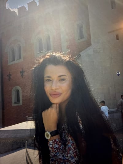 The photograph captures a woman with long, dark hair, resting her chin on her hand. She is wearing a patterned shirt and a watch on her left wrist. Behind her, a historic brick building with arched windows and crenellated battlements is visible. Sunlight streams from the upper right, casting a glow on the scene. In the background, two children and an adult are present, suggesting a public outdoor setting. The overall impression is that of a portrait taken on a sunny day, with a backdrop of ancient architecture. The specific location of Kozlovets, Bulgaria is not discernible from the image itself.