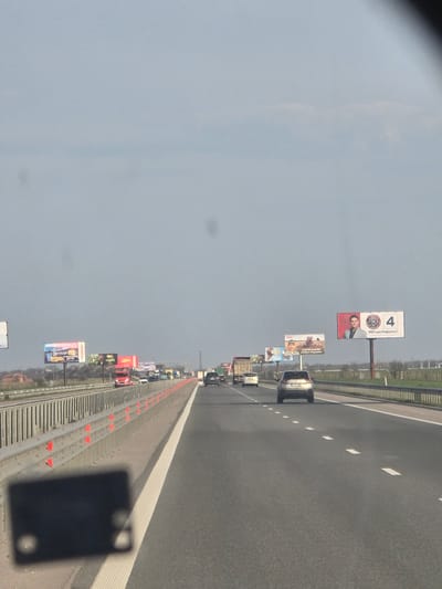 The image displays a daytime highway scene near Opalchenets, Bulgaria, observed from the interior of a forward-moving vehicle under a uniform light gray, overcast sky. The asphalt highway features at least two visible lanes of travel, clearly delineated by white dashed and solid lane markings. A dark, out-of-focus object, consistent with a dashboard reflection, occupies the bottom-left quadrant.
To the left, a metal guardrail with vertical elements and a secondary upper rail separates the highway from a dirt shoulder and distant green fields, beyond which several residential or commercial structures are partially visible. Orange reflective markers are affixed to the base of the guardrail. To the right, open green fields parallel the highway.
Traffic consists of several vehicles in motion. Directly ahead in the right lane is a silver SUV with partially visible Bulgarian license plate characters "34 98." Further ahead in the left lane is a white sedan, followed by a red semi-truck cab. Additional cars and a brown semi-truck are visible further down the road in both lanes, indicating moderate traffic flow.
Numerous billboards are positioned along both sides of the highway. On the left, one billboard displays "HESBURGER" branding with images of burgers and text in Cyrillic, including "СУПЕР ЦЕНА!" (Super Price!) and prices in Bulgarian Lev. Another billboard on the left features a man with a mustache, a tractor, and "АГРИТЕХМАШИНИ" (AgriTechMachines) text. On the right, a prominent billboard presents a man in a suit alongside the number "4" and a stylized symbol, with the Cyrillic text "МЕЧ ИЛИ МАФИЯТА?" (Sword or Mafia?), indicative of a political advertisement. Dust specks are visible in the upper sky portion of the image.