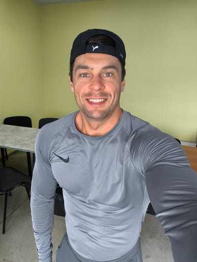 A man wearing a black baseball cap and a grey long-sleeved athletic shirt is taking a selfie in a room with light green walls. He is smiling and has short brown hair. In the background, there is a long grey table with two black chairs on one side, and a wooden desk on the other side of the room. The floor appears to be concrete. This scene is located in Molokovo, Russia.
