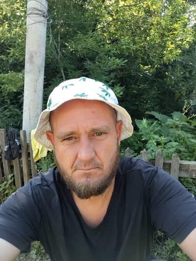 A man with a beard and a bucket hat is taking a selfie outdoors. He is wearing a dark t-shirt. Behind him, there is a wooden fence, a utility pole, and lush green foliage. The man is positioned centrally in the frame, looking directly at the camera with a neutral expression. The setting appears to be rural or suburban, given the presence of the fence and the natural vegetation.