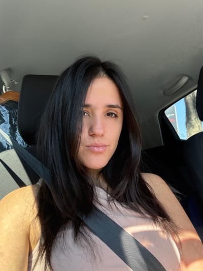 A woman with long, dark hair poses for a selfie inside a car. She wears a beige top and a black seatbelt diagonally across her chest. Her skin has a warm tone, and she looks directly at the camera with a slight smile. Sunlight streams through the car's window, creating highlights in her hair and shadows on her clothes. The car's interior is visible, with a light gray ceiling and dark gray seats. A glimpse of the outside sky is visible through the sunroof.