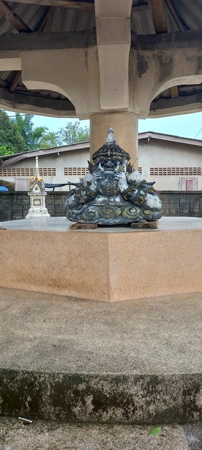 The scene is an outdoor shot in Ban Thung, Thailand, featuring a cement statue of a deity placed on a cement pedestal in front of a building. The statue, weathered and aged, has intricate carvings with gold detailing. Behind the statue is a building with rectangular windows. To the left of the statue is an architectural structure with a roof. The environment consists of a textured, cement surface with mossy patches.