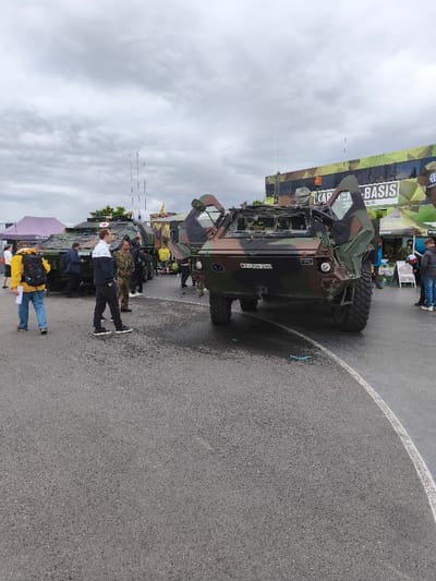 This image depicts an outdoor military display event in Hohenstein-Ernstthal, Germany, under an overcast sky. The foreground and midground feature dark asphalt pavement with a prominent curved white line extending from the bottom right. Centrally positioned is a multi-camouflaged German Bundeswehr Fennek armored reconnaissance vehicle, oriented towards the viewer. Its front armored doors are open, revealing the internal compartment. The vehicle's front displays the license plate "Y-304 240." To its left, partially obscured by individuals, stands another multi-camouflaged military vehicle, consistent with a GTK Boxer. In the right background, a large camouflaged Bundeswehr promotional truck or bus is visible, featuring text that includes "KARRIERE BEI DER BUNDESWEHR" and the Bundeswehr insignia, adjacent to a green display tent. A purple tent is situated in the far left background. Approximately 10-15 individuals are present, consisting of civilians and at least one uniformed Bundeswehr soldier. Civilians include a person in a yellow jacket with a backpack on the left and another in a white and dark blue jacket walking in the midground. All individuals appear to be observing the military vehicles and displays. Patches of moisture are discernible on the asphalt beneath the front of the Fennek vehicle.