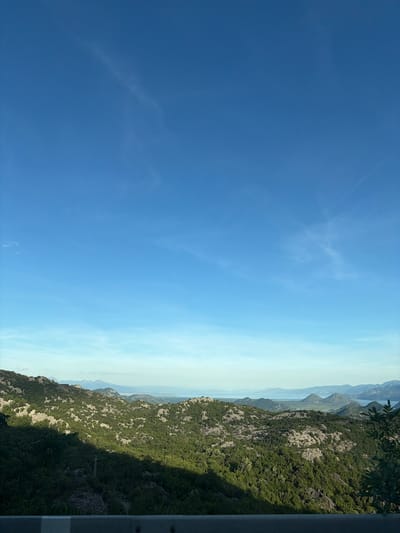 The image displays a panoramic view of a mountainous, wooded landscape under a clear blue sky with wispy clouds. In the foreground, a concrete barrier with white stripes suggests a roadside viewpoint. The middle ground is dominated by lush green trees and rocky terrain, with a prominent shadow cast across the landscape, indicating the time of day. In the distance, a body of water, possibly a lake or sea, is visible, surrounded by more rolling hills and mountains that fade into the horizon. There are no people or animals visible in the scene. The location is identified as Gornja Brova, Montenegro.
