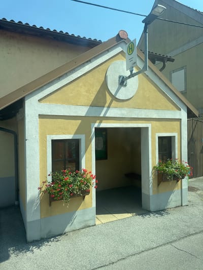 The image shows a small yellow bus stop in Wolfsthal, Austria. The bus stop is shaped like a miniature house with a pitched roof. It has a white trim around the edges and is decorated with flower boxes containing red geraniums on either side of the entrance. A bus stop sign with a green circle and an "H" symbol is mounted on a pole to the left of the entrance, indicating the stop's name as "Wolfsthal Kirche" with "VOR" written below it. The interior of the bus stop has a wooden bench. The ground in front of the bus stop is paved with a light gray speckled asphalt. The sky is clear and blue.