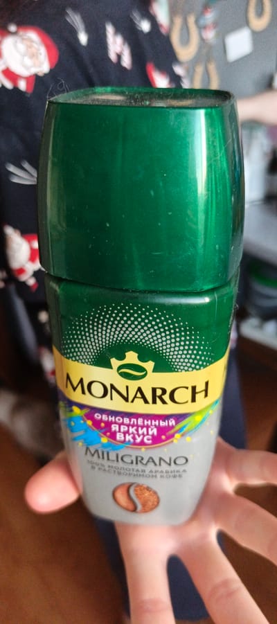 An upside-down, clear plastic container of Monarch Miligrano coffee is being held by a person's hand in Tikhvin, Russia. The container has a green lid and a label with yellow, purple, and blue accents. The label features the Monarch logo, the words "ОБНОВЛЕННЫЙ ЯРКИЙ ВКУС" (Updated Bright Taste), and "MILIGRANO." A close-up image of coffee beans is also visible on the label. The background of the image is blurred but appears to be a dark, patterned fabric with a Christmas theme, featuring images of Santa Claus and other festive elements. The lighting suggests an indoor environment.