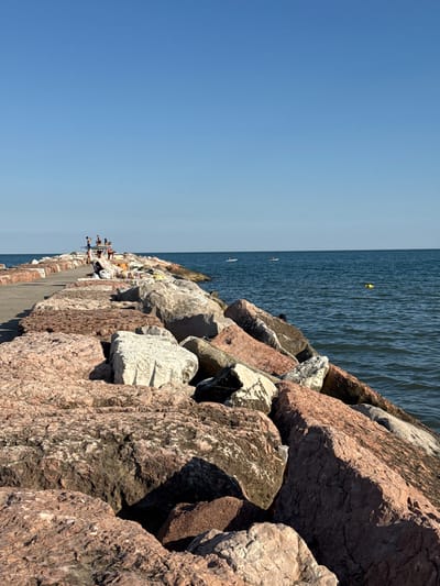 The image depicts a breakwater made of large, rough, reddish-brown and white rocks leading into a calm, deep blue sea under a clear, light blue sky. On the breakwater, in the distance, a small group of people are visible. Some are standing and appear to be interacting, while others are seated or lying down on towels. Further out on the water, there are a few individuals on paddleboards, and a yellow buoy is floating. The overall scene suggests a sunny, recreational day at a coastal location in Eraclea, Italy.