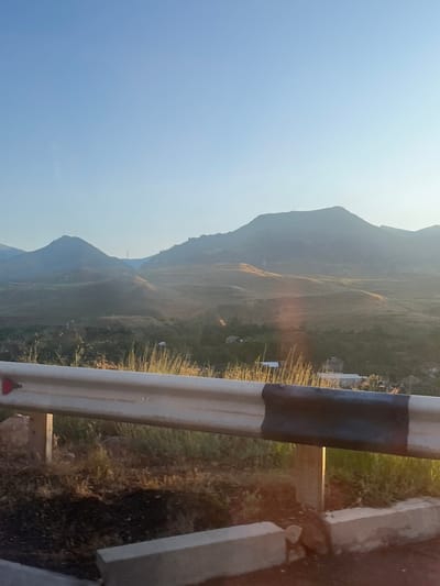 The image captures a scenic view of rolling hills and mountains under a clear blue sky, likely taken from the side of a road. A white and black metal guardrail is prominently positioned in the foreground, with some dry grass and vegetation growing around it. In the mid-ground, small buildings and scattered trees are visible amongst the undulating terrain. The landscape is bathed in a warm, hazy light, suggesting it might be early morning or late afternoon. The overall impression is one of a rural, mountainous environment. The location is identified as being in the city of Arpi, Armenia. There are no people or animals visible in the image.