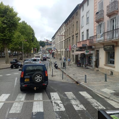The image depicts a street scene in Foix, France, likely on a cloudy day. In the foreground, a dark blue Land Rover Discovery is stopped at a pedestrian crossing. The vehicle has a spare tire mounted on the rear, a yellow license plate that reads "1316 GS 09," and the "DISCOVERY" badging is visible. The pedestrian crossing is marked with white stripes on the asphalt.
To the right of the street, a paved sidewalk runs alongside a row of multi-story buildings. The buildings have a mix of architectural styles, some with ornate balconies and shutters, and others appearing more utilitarian. Various storefronts are visible at street level, including "CENTURY 21 L'Agence," "PMU express," and what appears to be a newsstand or tobacco shop labeled "PRESSE." Bicycles are parked on the sidewalk near one of the buildings.
On the street, an ATV with two riders wearing helmets is moving away from the viewer. Further down the street, other vehicles, including cars and a camper trailer, are visible in traffic. A construction crane is also visible in the background, suggesting ongoing development or renovation in the city. Trees line the left side of the street, providing shade. The overall impression is a typical urban street in a French town.