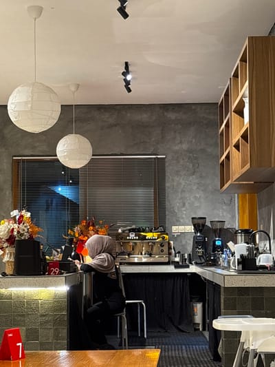 An interior view depicts a contemporary coffee shop located in Patrang, Indonesia. The scene is characterized by a grey concrete-effect wall, a white ceiling, and a dark floor. Two large white spherical paper lanterns and multiple black track spotlights illuminate the area. A window with horizontal blinds is visible in the background, revealing a blue ambient light outside.

A woman, wearing a light brown hijab and dark clothing, is seated on a white stool behind a counter in the midground. She is leaning forward, engaged with items on the countertop. The counter, which has a speckled grey surface and a greenish-grey tiled front on the left, extends across the image. On the left side of the counter, a dark speaker, a white basket with artificial flowers (red, orange, yellow, white), and a red "29" table sign are visible. To the right of the woman, a multi-group espresso machine with gold and chrome finishes is positioned. Further right, the counter features two black coffee grinders, a black gooseneck kettle, and various barista tools and containers. The lower section of the counter on the right is covered with black fabric, concealing a visible white trash bin.

An open wooden shelving unit with a grid design is mounted on the upper right wall. In the foreground, a portion of a wooden table is visible, adorned with a red triangular sign displaying "TABLE 7". A white high chair is partially visible on the far right, near the counter.