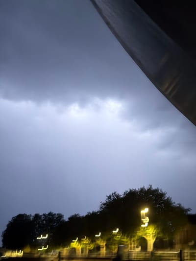 This is a photograph taken at dusk in Charnay-lès-Mâcon, France, facing upwards towards a clouded sky with a large, dark, angled structure in the upper right corner, possibly a building or overhang. The lower portion of the image shows a silhouetted line of trees against the dim sky, with blurred yellow lights from streetlamps or windows creating streaks of light along the bottom. The overall atmosphere is moody and indistinct due to the low light and motion blur. There are no people or identifiable actions visible in the image.