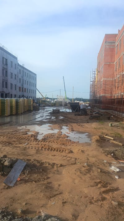 The image depicts a construction site in the Babovichi rural council, Belarus. Two large, multi-story buildings are under construction, one on the left with a grey facade and visible window openings, and another on the right constructed of red brick. Piles of building materials, including what appears to be stacked pallets of packaged items on the left and scaffolding surrounding the brick building on the right, are visible. The ground is a mix of wet, muddy soil with tire tracks from vehicles, and dry, sandy areas. Several cranes are present on the site, including a tall green crane in the background. Construction debris and assorted building materials are scattered throughout the foreground. The sky is overcast with pale blue and white clouds. There are no people visible in the frame.