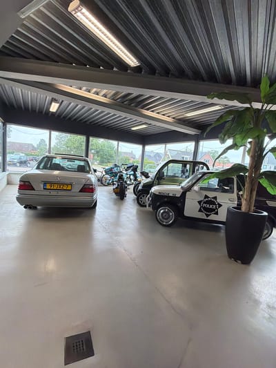 The image shows the interior of a commercial building, likely a showroom or garage, located in Varsseveld, Netherlands. The space has a polished concrete floor and a ribbed metal ceiling with fluorescent lighting fixtures. Large windows line the back wall, offering a view of a street with houses and trees, suggesting an outdoor environment under an overcast sky.
Inside, the main focus is a collection of vehicles. In the foreground and to the left, a silver Mercedes-Benz sedan is parked. To the right of the sedan, there are several motorcycles, including one with a blue fairing. Further to the right, two distinct small vehicles are positioned: a dark green, utilitarian-looking vehicle, and a black and white vehicle designed to resemble a police car with "RESCUE 112" and "UNITED STATES POLICE VARSSEVELD" markings. A large potted plant with green foliage stands to the right of the police-style vehicle, partially obscuring a dark-colored vehicle behind it.
There are no people visible in the image. The overall scene depicts a well-lit indoor display of vintage and custom vehicles within a commercial property in Varsseveld.