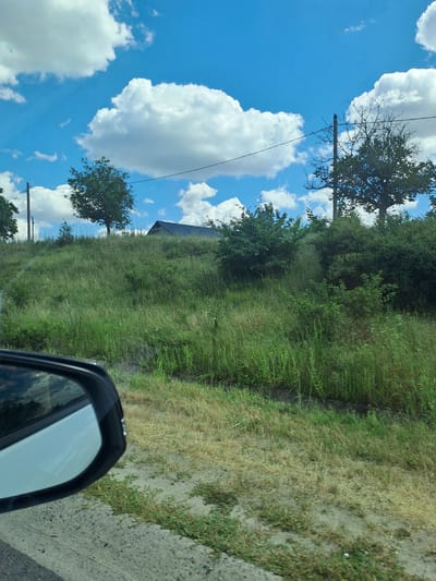 A side view captured from a vehicle shows a bright, sunny day with a vivid blue sky dotted with fluffy white clouds. The scene is dominated by a grassy incline covered in lush green vegetation, interspersed with dry, yellow grass and patches of bare earth. A dark-colored side mirror is prominently featured in the foreground, reflecting the sky and surrounding greenery. In the mid-ground, several trees of varying sizes and foliage density are visible, with a building with a dark, sloped roof situated on top of the hill. A utility pole with wires stretches across the upper portion of the image, contributing to the rural or semi-rural ambiance. The image depicts a landscape in or near Budești, Moldova.