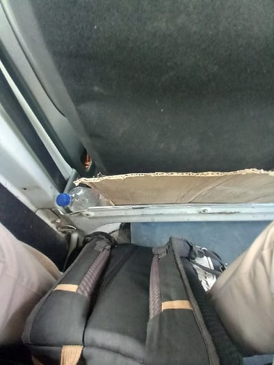 The image shows the interior of a vehicle, possibly a bus or van. The vehicle is in the city of Zaghouan, Tunisia, at the coordinates 36.4 latitude and 10.148 longitude. The image is taken from inside the vehicle, looking towards the back. A large, black backpack is visible in the back, resting against the back seat. Part of a brown garment can also be seen next to the backpack. A partially visible plastic water bottle is lying on a shelf above the backpack. The seat is beige in color and worn.  The overall lighting is dim, suggesting an interior environment. There are no people visible in the scene.