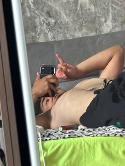 This is a mirrored, upside-down, close-up shot of a person in a bedroom. The person is lying down, with their torso and arm visible. They are wearing black shorts and appear to be tan. Their right arm is bent, and they are holding a smartphone in their right hand, taking a selfie. Their left hand is raised, and their index finger is extended, possibly pointing or gesturing. The person's face is visible in the mirror, and they have an engaged expression.
In the background, a grey, textured surface, likely a floor or wall, is visible. To the right of the person, there is a bright green fabric, possibly a blanket or clothing. A black and white patterned fabric is also visible between the person and the green fabric.
The lighting suggests it's daytime, though the specific time and weather are not discernible. The scene appears to be an indoor setting, most likely a bedroom, given the presence of bedding-like fabrics. No text is visible in the image. The overall mood is casual and self-aware, typical of a mirror selfie.