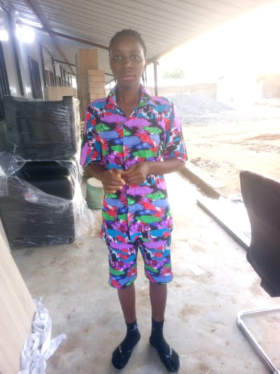 A young Black woman stands in what appears to be an outdoor or semi-enclosed workshop or storage area, possibly in Indo, Nigeria. She is wearing a colorful, patterned short-sleeved shirt and matching shorts. Her hands are clasped in front of her. To her left are stacks of large, black objects wrapped in plastic. In the background, there are piles of gray gravel and some brick structures. The ground is concrete, with some dirt and debris. The lighting suggests it is daytime, with strong sunlight filtering in from the side.