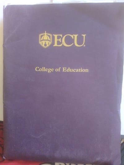 The image is of the cover of a document, likely a brochure or report, from the College of Education at ECU. The cover is a dark purple color with gold lettering. In the upper center of the cover is a gold emblem, which appears to be a stylized shield with a crown or spire on top. To the right of the emblem, in large gold capital letters, is the text "ECU". Below this, in smaller gold lettering, is the text "College of Education". The cover appears to be slightly worn, with some faint scuff marks visible. The lighting is even, suggesting indoor illumination. There is no indication of the time of day or weather conditions. The location context provided, Magajin Gari, Nigeria, does not have any direct visual cues within the image itself to confirm or deny this location. The overall impression is formal and academic.