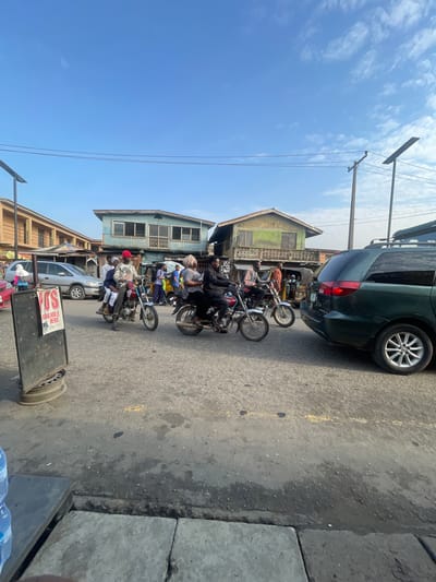 The image depicts a street scene in Akowonjo, Nigeria. In the foreground, a dark-colored van is parked on the right side of the road. Several motorcycles are in motion. One motorcycle, ridden by a person wearing a red cap, carries three passengers. Another motorcycle is ridden by a person in a patterned shirt, with a passenger behind them. A third motorcycle is partially visible on the left. In the background, buildings with a weathered appearance line the street. A silver van is parked to the left, and several people are walking. The road surface appears to be asphalt, with some signs of wear and tear. The sky is clear and blue. A street lamp is visible on the left side of the image.