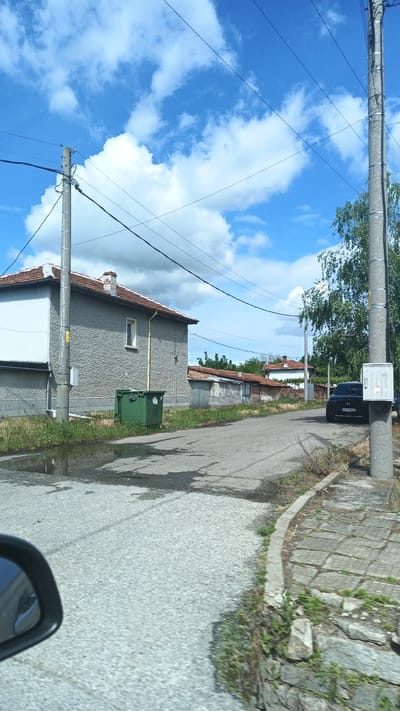 A street in Boliartsi, Bulgaria. On the left is a stone building with a red tile roof. Next to the building is a green dumpster. A puddle is on the road in front of the building. Further down the road is a black car parked next to a telephone pole. The sky is blue with clouds. Telephone wires are visible above the street. A car's side mirror is visible in the foreground.