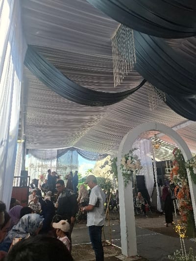 The image shows a wedding ceremony taking place under a white and blue canopy tent in Bojongsoang, Indonesia (latitude: -6.968, longitude: 107.651). The ceremony is held outdoors, with a white archway decorated with flowers marking the focal point. People are gathered around the archway, including a man in a white shirt and blue jeans standing in the center, and a woman wearing a white hijab. Several other people are visible in the background, some sitting and some standing. The canopy tent is adorned with white and blue fabric drapes and hanging decorations. The scene is bright and lively, with a festive atmosphere.