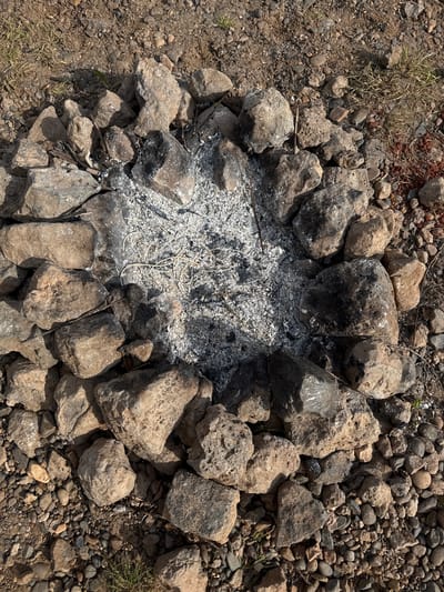 The image shows a circular fire pit constructed from roughly hewn rocks and stones. The interior of the pit contains ash and charred remnants of burned material. The surrounding ground is a mixture of soil, gravel, small stones, and sparse vegetation. The location is unspecified, and there is no indication of human presence in the image. The scene depicts a fire pit in a natural or semi-natural environment. There is no evidence to indicate the location as Povarenki, Russia.