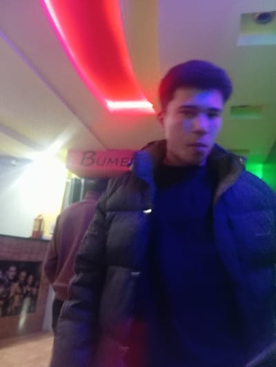 An indoor scene in Takhtakuprik, Uzbekistan, features a young man centered in the foreground, facing the camera. He has dark hair and wears a dark puffer jacket over a dark shirt. Blue light illuminates the right side of his face and parts of his jacket. Behind him, to the left, another individual is partially visible from the back, wearing a reddish-brown garment. The environment is characterized by artificial colored lighting, with a prominent red curved light fixture on the ceiling and a green light source on the right side of the frame. A sign partially displaying the letters "BUME" in red is visible in the mid-ground. To the left, a light-colored wall features a dark mantelpiece or counter structure, above which a red cylindrical object, possibly a fire extinguisher, is mounted. Below the mantelpiece, a framed image depicting a group of people is visible. The floor appears light-colored and uniform. No direct interaction between the individuals is evident.