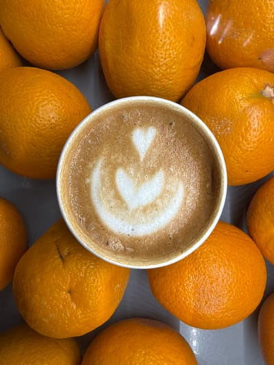 This overhead shot features a white cup of coffee with intricate latte art, centrally placed and surrounded by numerous bright orange citrus fruits. The coffee, likely a cappuccino or latte, displays a well-executed white foam pattern resembling a heart or stylized leaf, set against the warm brown of the beverage. Small bubbles are visible around the edge of the foam.
The cup is nestled among several ripe oranges, which fill the rest of the frame. These oranges have a vibrant, uniformly orange color and distinctly textured, bumpy skins. Some oranges show small, dark marks or remnants of stems. They are arranged closely together, creating a dense backdrop for the coffee cup.
The overall scene is a still-life composition, suggesting a moment of pause or a casual arrangement of breakfast or snack items. There is no explicit activity or event taking place. The lighting is even and bright, highlighting the textures of both the coffee foam and the orange peels. No specific time of day, weather, or emotions are discernible from the image. There is no visible text. Although the image is from Вёшки, Russia, no specific visual cues within the photograph indicate this location.