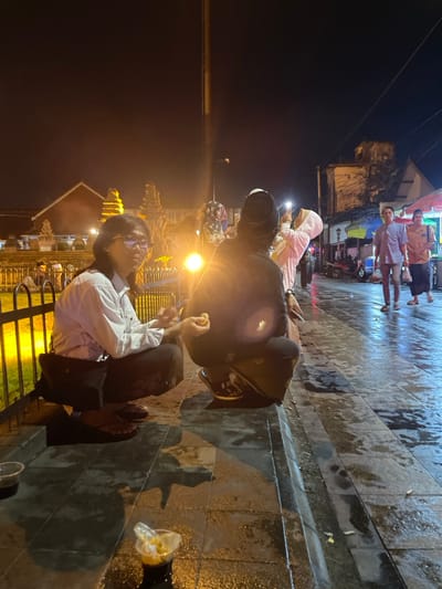 The image displays a nighttime street scene in Kauman, Indonesia, characterized by a dark sky and wet pavement reflecting artificial light. In the foreground, two individuals are squatting on a tiled sidewalk. The person on the left, with long dark hair and eyeglasses, wears a white long-sleeved shirt, dark pants, and flip-flops, holding a small, light-colored food item in their right hand while looking towards the camera. A black shoulder bag is slung over their left shoulder. The second individual, positioned to their right and facing away from the camera, wears a dark hooded garment, dark pants, and sneakers, holding a similar light-colored food item in their right hand. A dark cap covers their head. On the pavement near them, two clear plastic cups are visible; one contains a dark liquid with ice, and the other holds light-colored contents and a plastic wrapper.

Behind the squatting individuals, a metal fence separates the sidewalk from an illuminated area featuring traditional Indonesian architectural structures with distinct layered roofs and golden-yellow ornamentation, backed by a red-roofed building. A tall, dark pole ascends vertically from the mid-ground. Further to the right, a third individual, wearing a light-colored head covering (hijab), is partially visible, holding a smartphone with its flashlight activated.

In the background, a wet asphalt street runs parallel to the sidewalk, reflecting ambient light. Two men are walking away from the camera; one wears a light purple shirt and light pants, the other a tan shirt and dark pants. The street is lined with multi-story buildings displaying aged facades and numerous electrical wires, alongside market stalls with colorful awnings.