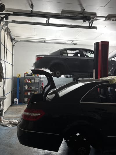 This media file depicts an indoor scene within a car repair shop. The primary subjects are two dark-colored cars positioned on a car lift. One car is partially raised, allowing access to its underside, while the other car is positioned in front of it. Various tools and equipment associated with automotive repair are visible, including shelves stocked with containers and bottles, a blue mechanic's creeper, and chains likely part of the garage door mechanism. The setting appears to be a well-lit garage with white paneled walls. The lighting suggests it is daytime, as is typical for a business operating hours. There is no visible weather information as the scene is indoors. There is no visible text in the image. The overall mood is functional and professional, indicative of a working environment.