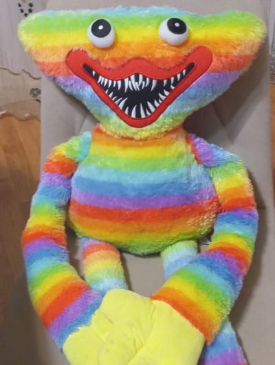 This is a photograph of a stuffed toy, likely a plush doll, with a rainbow-colored body. The toy has a long, segmented body with arms and legs. Its head is flat and triangular, with two large, googly eyes and a wide, red mouth filled with sharp, black and white teeth. The body, arms, and legs are striped with the colors of the rainbow: red, orange, yellow, green, blue, and purple. The toy's arms and legs are bent, giving it a somewhat contorted or stretched appearance. One of its limbs ends in a bright yellow section. The toy is lying on a textured surface, possibly a sofa or carpet, with a blurred background that suggests an indoor setting. The lighting appears to be artificial, and the image is taken from a slightly elevated, overhead angle. No people are visible in the image. The setting does not provide specific cues to determine the time of day or weather.