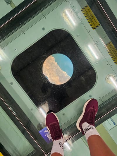This image shows a first-person perspective of a person standing on a glass floor with a circular opening that reveals a view of rocky terrain and the sky below. The person is wearing burgundy athletic shoes with white soles and white socks with black text. The glass floor is part of a structure with a teal-colored frame. Yellow warning labels with black stripes are visible on the frame, and there are ventilation grates. A blue QR code sticker with the text "SPACE VIEW" is also visible on the glass floor. The location appears to be an observation deck or a similar attraction. The provided information indicates this is the city of Mukilteo, United States.