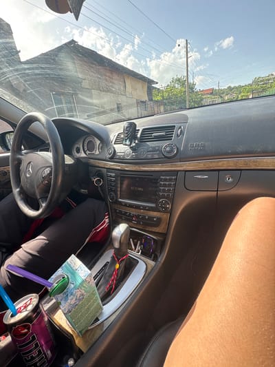 The image shows the interior of a Mercedes-Benz car, focusing on the dashboard, steering wheel, and center console. Visible items include the car's radio displaying "87.5 MHz", various buttons and knobs on the dashboard, and the steering wheel with the Mercedes-Benz logo. In the center console, there's a can of "HELL" energy drink, a juice box, and several straws. Part of a person's leg is visible on the right side of the image, near the car door.

Looking through the windshield, there's a view of a building and utility lines, indicating an urban or suburban environment. Since the city of Rila, Bulgaria, is mentioned, it can be inferred that the image was taken somewhere in Rila. No specific location within Rila can be pinpointed from the image alone.