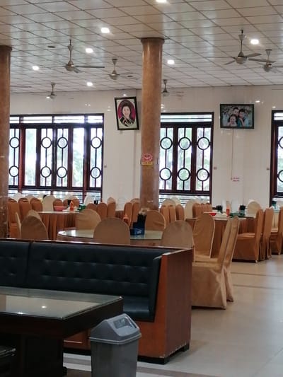 The image shows the interior of what appears to be a dining hall or restaurant in Chu Văn An Ward, Vietnam. The space is large, with numerous round tables covered in cloths. Chairs, also draped in fabric, surround each table. The seating area extends into the distance, indicating a substantial capacity. The furniture has an older, traditional style.

Visible are large windows with decorative circular patterns, allowing natural light into the room. Two framed pictures hang on the walls, one showing a person, and the other showing a family. Above, the ceiling features a grid pattern with multiple ceiling fans.

In the foreground is a dark-colored booth or sofa. A glass-topped table sits in front of it. Next to the table, there is a trash can. The overall impression is that of a functional, moderately decorated space, likely used for large gatherings or events.
