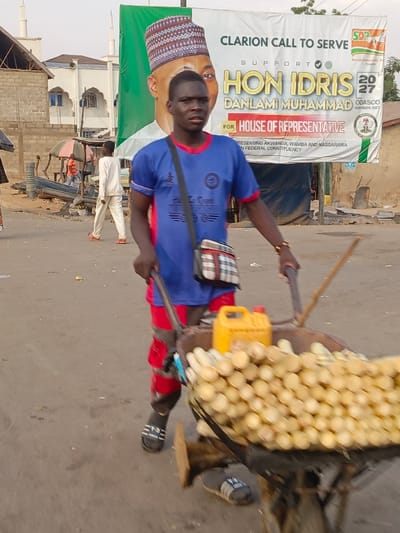 In an outdoor scene, a man pulls a wheelbarrow filled with what appear to be peeled sugar canes. He wears a blue T-shirt, red shorts, and sandals. A banner in the background advertises support for Hon. Idris Danlami Muhammad for House of Representatives, referencing the Social Democratic Party (SDP) and representing Akwanga, Wamba, and Nassarawa in a federal constituency. A mosque is also visible in the background.