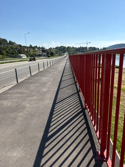 This image shows a view of a bridge with a red railing and a paved walkway. The sun is shining, casting long shadows from the railing onto the walkway. In the distance, there are trees, houses, a body of water, and a construction crane. The sky is clear and blue. There are a few people walking on the walkway, and what appear to be bicycles parked near the railing. The scene is well-lit and appears to be a sunny day. The image is taken from a high angle, looking down the length of the bridge. The location context is Gródek nad Dunajcem, Poland. No visible text is present in the image.