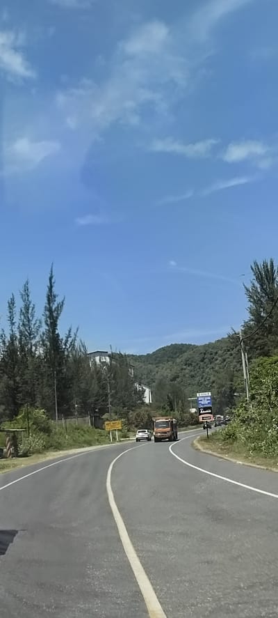 A vertical, eye-level, medium shot captures a road curving to the left and uphill with vehicles. The road is paved with asphalt and marked with a solid white line on the right and a dashed white line on the left. In the foreground, the road dominates the lower half of the frame. On the left side of the road, there are some sparse, tall trees with thin needles and a short, brown wooden structure. On the right side, the road is bordered by lush green vegetation and trees.
Further down the road, a white passenger car is visible, followed by an orange flatbed truck. Beyond these vehicles, the road continues to curve uphill. On the right side of the road, a blue billboard with white text is placed next to a green bush. In the distance, a multi-story building with a white exterior and a gray roof is situated on a hillside. The hillside is covered with dense green foliage and trees, creating a natural backdrop. The sky above is a clear, bright blue with scattered white clouds. The overall scene depicts a road in a natural, semi-urban environment, likely in Lamkruet, Indonesia, with a mix of man-made structures and abundant greenery.