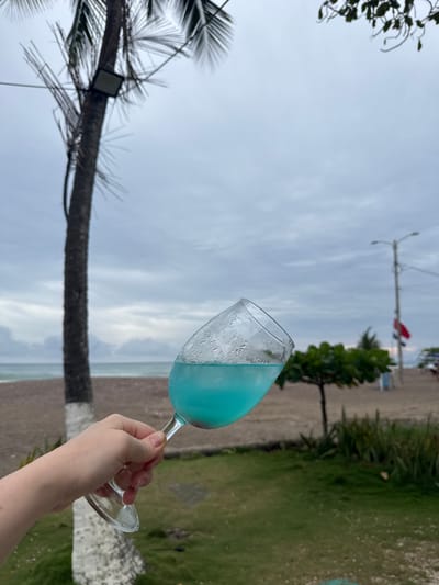 A person's hand holds a clear glass with a blue beverage and ice. The glass is tilted, obscuring some of the liquid. The background features a beach with dark sand, palm trees, and a cloudy sky. A street lamp and two flags are visible on the right side of the image. This scene is set in Jacó, Costa Rica.