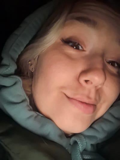 A close-up selfie of a young woman with fair skin, light blonde hair, and brown eyes wearing a hooded sweatshirt. The hood is up, framing her face. She has dark eyelashes and a subtle smile. The lighting is dim, casting shadows on the side of her face. The background is dark and out of focus.