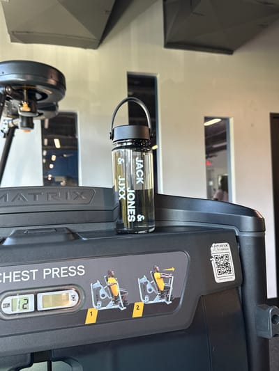 The image is a close-up shot of a black Matrix brand chest press exercise machine in a gym. A clear water bottle with a black lid and handle is positioned on top of the machine. White text on the bottle reads "JACK & JJXX JONES & JJXX". The machine has instructions for use displayed, including illustrations of a person performing the chest press exercise and a digital display showing "12". A QR code with the text "MATRIX ULTRA / CHEST PRESS" and "Scan for Instructional Video" is affixed to the right side of the machine. The background shows a gym environment with white walls, windows, and ceiling structures. The specific location in the City of Yonkers, United States of America, cannot be determined from the image.