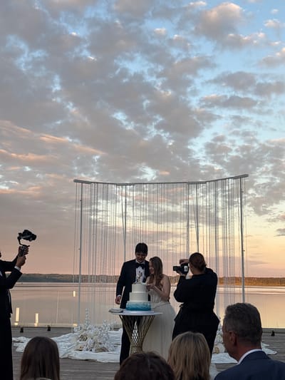 In Osipovo, Russia, a wedding reception takes place outdoors during sunset. A bride and groom stand behind a tiered cake, with the groom in a tuxedo and the bride in a white gown. The groom is holding a knife, about to cut the cake. A woman in a black top and pants films the couple with a camera. In the background, a decorative archway with hanging crystals forms a backdrop. A man holding a gimbal with a camera is also visible on the left side of the frame. Guests are seated in the foreground, observing the ceremony. The setting appears to be a waterfront location, with a body of water and a treeline visible in the distance. The sky is filled with dramatic clouds, illuminated by the warm hues of the setting sun.